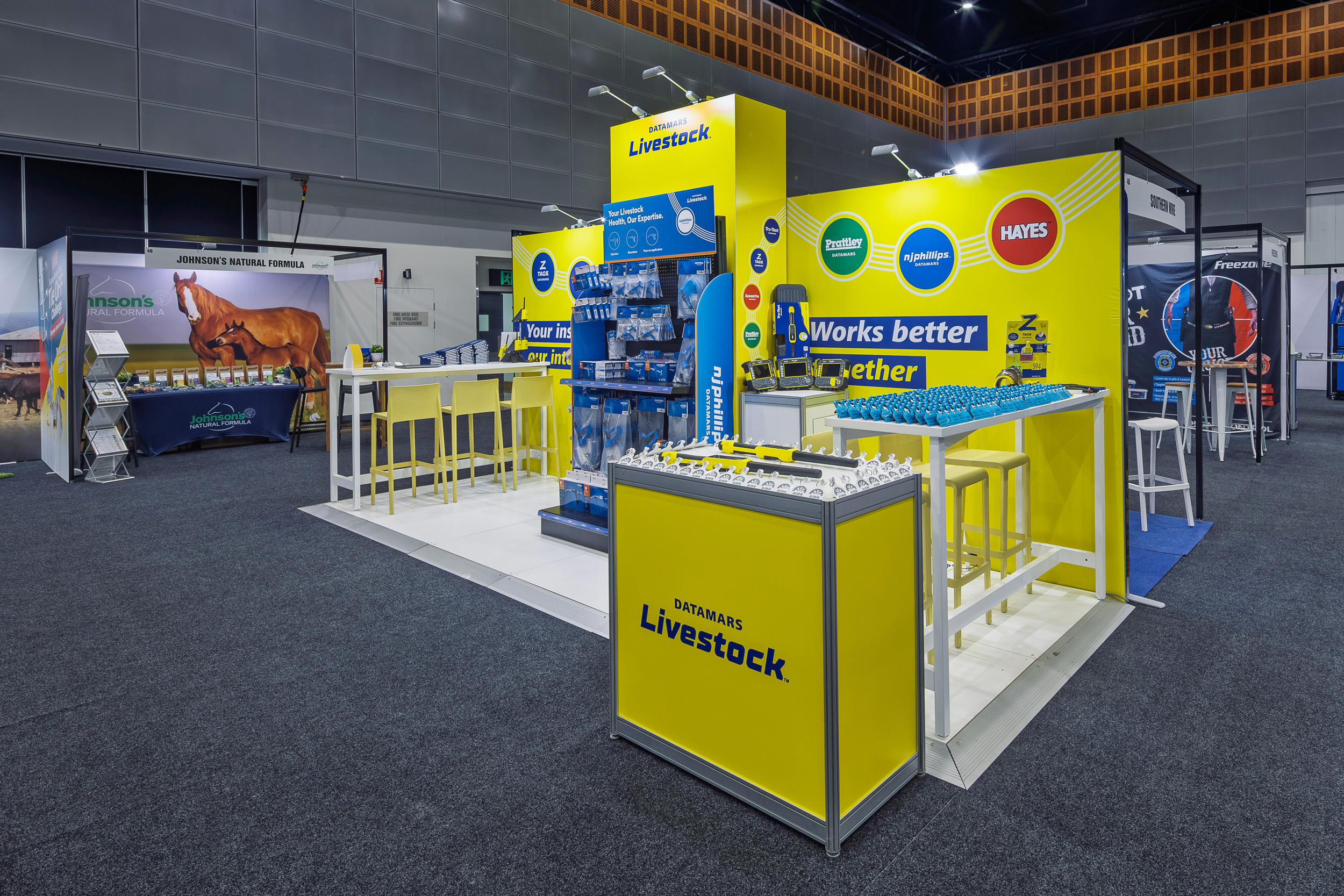 Livestock-themed exhibition booth with bright yellow displays, branded products, and informational signage. White stools and tables surround the area, set within a larger exhibition hall.