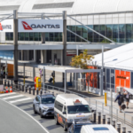 Exterior view of a Qantas terminal with people walking, a street with a white vehicle, and signage for Jetstar in a bright, clear setting.