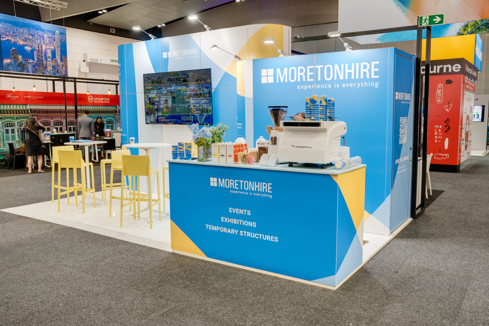 Trade show booth with blue branding for "Moreton Hire," featuring a counter, coffee machine, chairs, and a TV display. The text promotes events, exhibitions, and temporary structures.