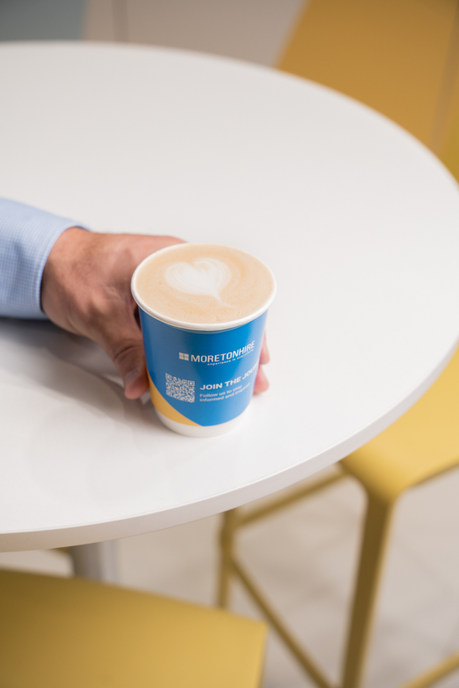 A person in a blue shirt holds a blue cup of coffee with latte art on a white round table, surrounded by yellow chairs.