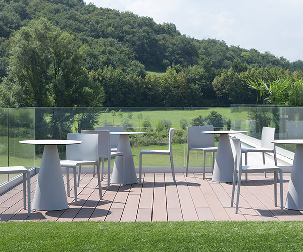 Outdoor patio with modern white tables and chairs on a wooden deck, overlooking lush green hills and trees. Clear glass railing borders the area.