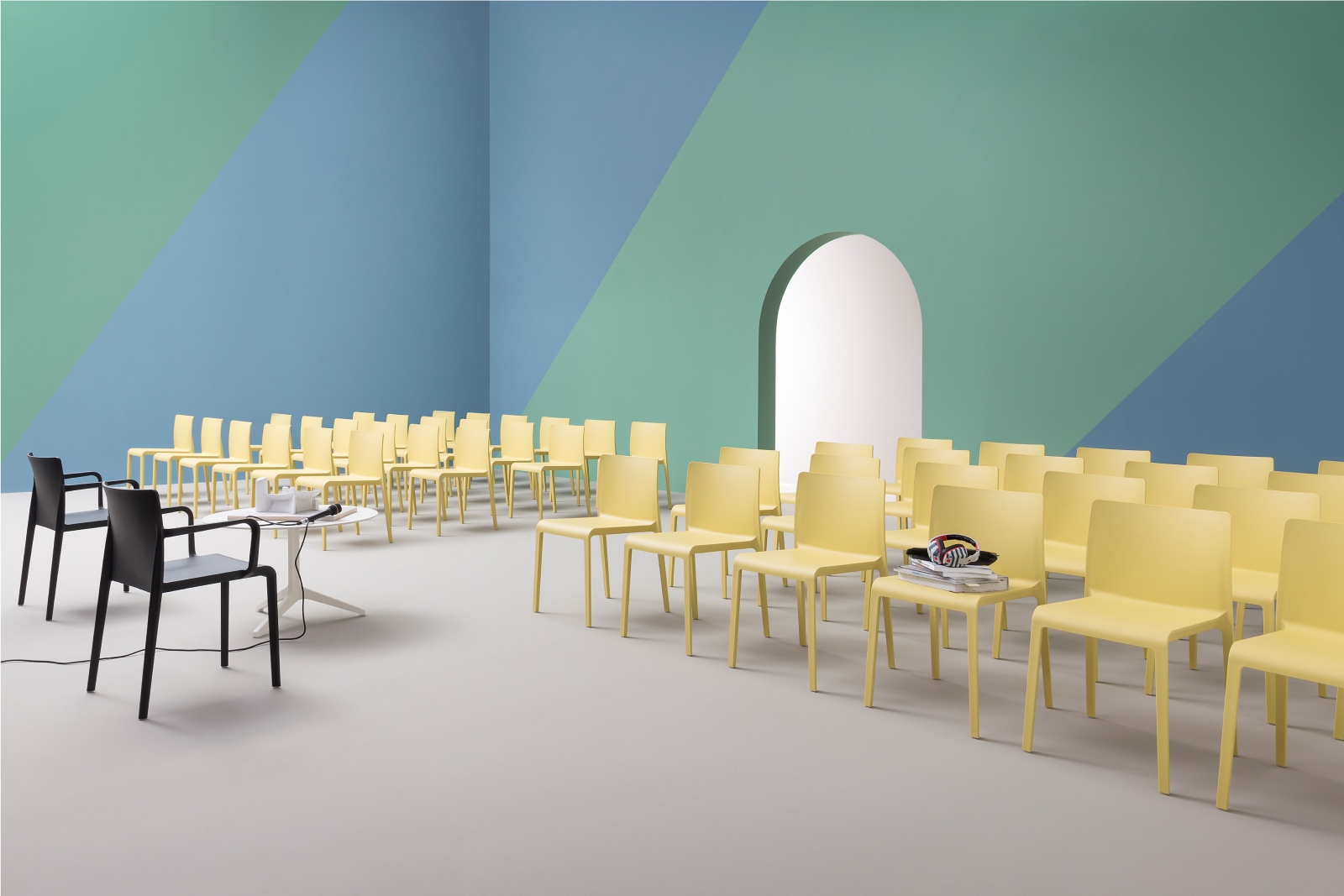 A room with rows of yellow chairs facing an archway, two black chairs with a small table in front, and a pastel blue and green wall.