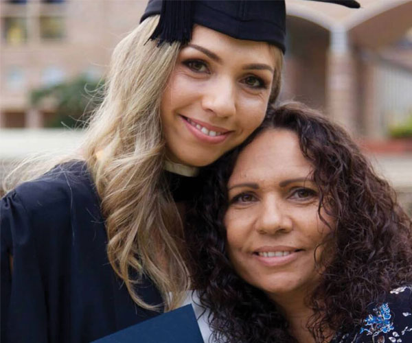 Two people sitting indoors at a table, wearing business attire and name badges. The woman is smiling, and the man beside her is looking at something on the table.