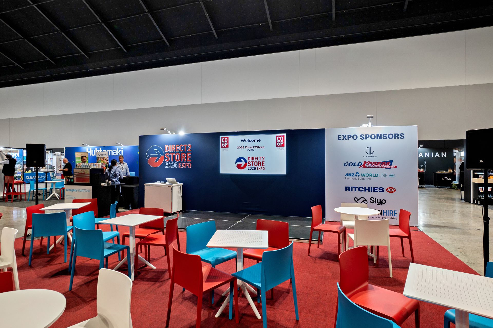 Trade-show floor with red, blue, and white chairs around white tables in front of a blue Expo booth reading Direct2Store 2026 Expo and a sponsor panel on the right.
