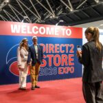 Couple posing for a photo at a large expo booth with 'WELCOME TO DIRECT2 STORE 2026 EXPO' text while a woman in black films them with her phone.