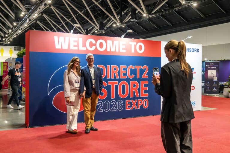 Couple posing for a photo at a large expo booth with 'WELCOME TO DIRECT2 STORE 2026 EXPO' text while a woman in black films them with her phone.