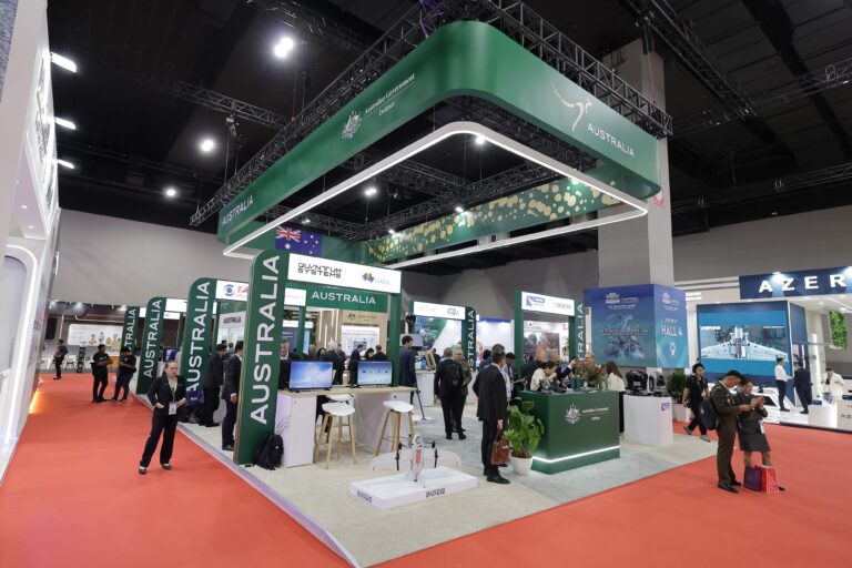 Wide view of an international expo hall featuring a large Australia booth with green overhead signage and many attendees exploring displays at counters and kiosks.