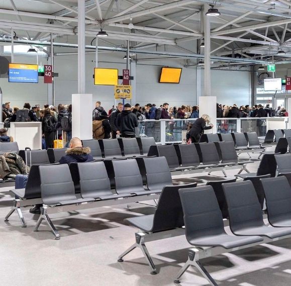 Busy airport terminal with many passengers lined up near counters, rows of gray seats in the foreground, and overhead signage in the background.