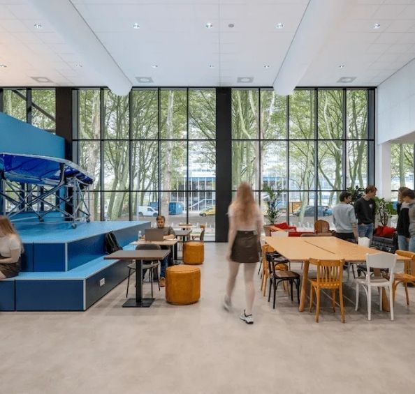 Open indoor common area with large floor-to-ceiling windows; people sit at tables, a blue multi-level stage is on the left, and a person walks toward the camera.