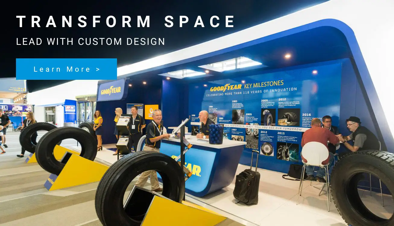 Trade show booth featuring a Goodyear display, large tires on yellow stands, and people talking near a blue information desk.