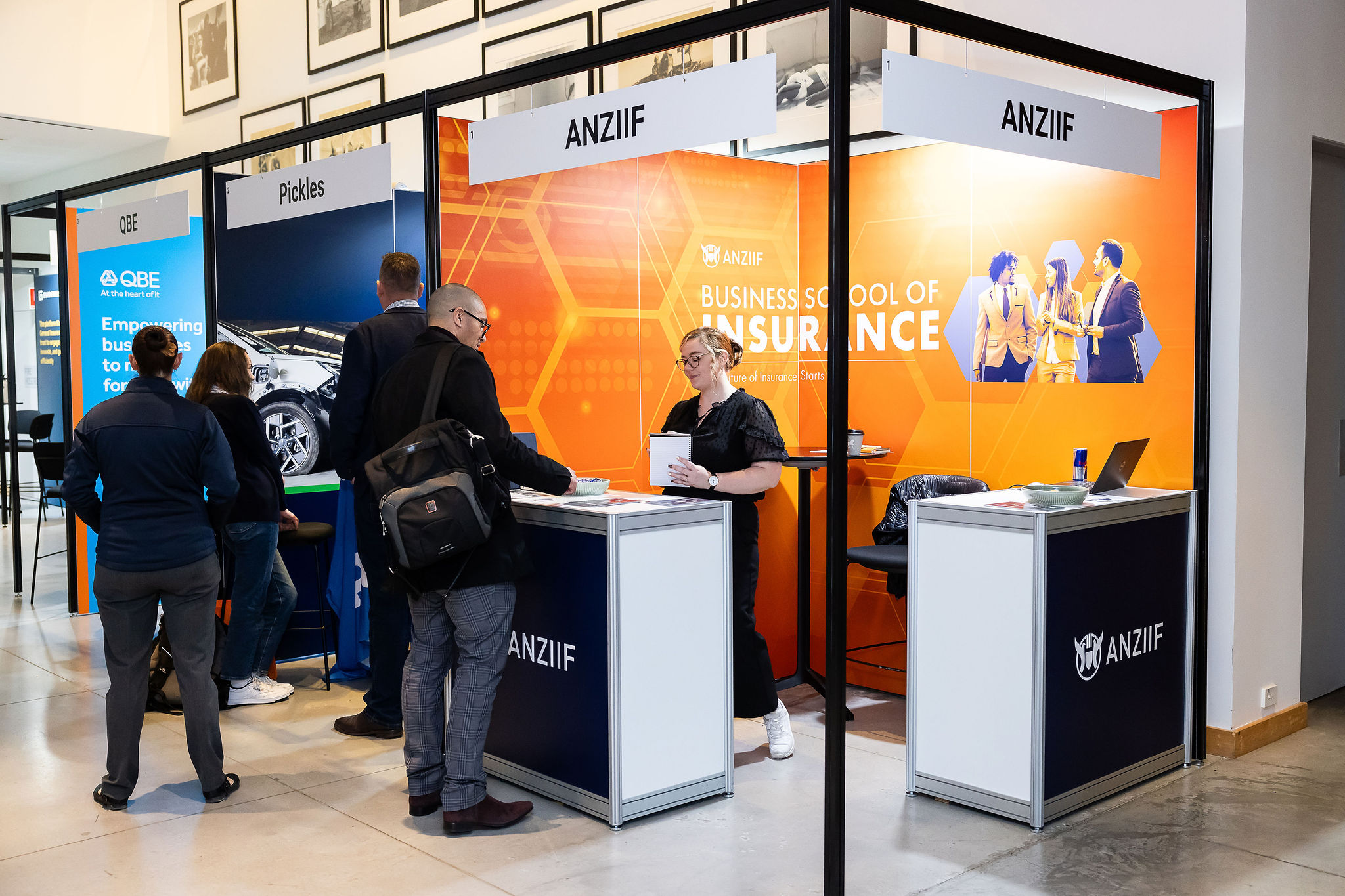 ANZIIF exhibition booth with orange graphics, two staff at a counter and attendees nearby in a meeting area.