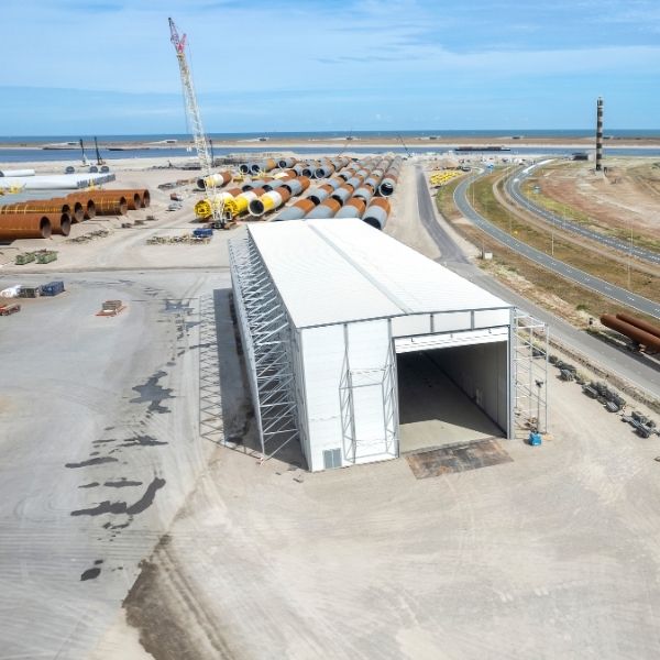 A large white temporary warehouse on a coastal construction site near the sea, with stacked steel pipes, a crane, and a curved road nearby.