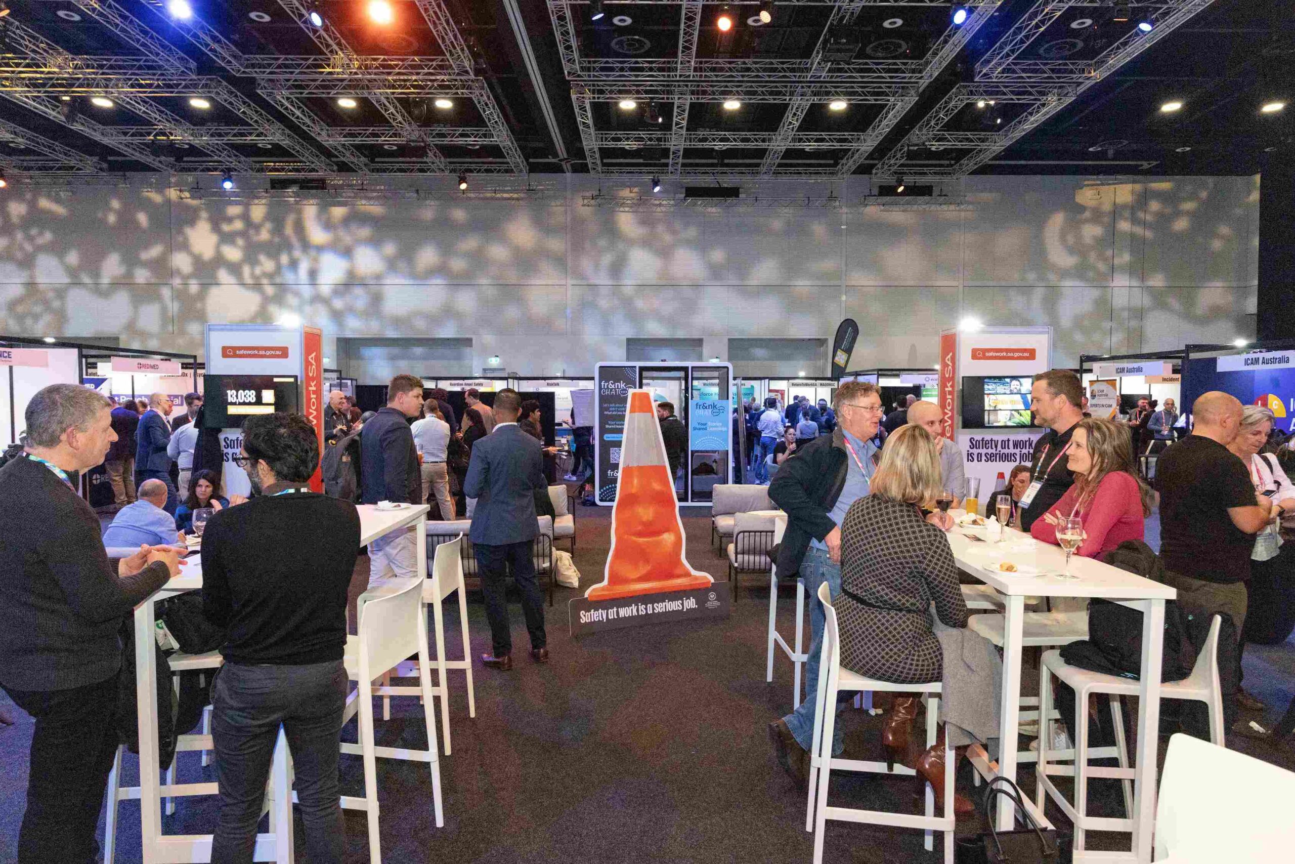 Busy conference expo hall with attendees around tables and vendor booths under bright overhead lighting.