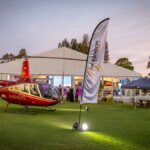 Red helicopter on a grassy field at an outdoor event with a white tent and banners in the background, lights illuminating the scene