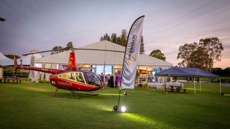 Red helicopter on a grassy field at an outdoor event with a white tent and banners in the background, lights illuminating the scene