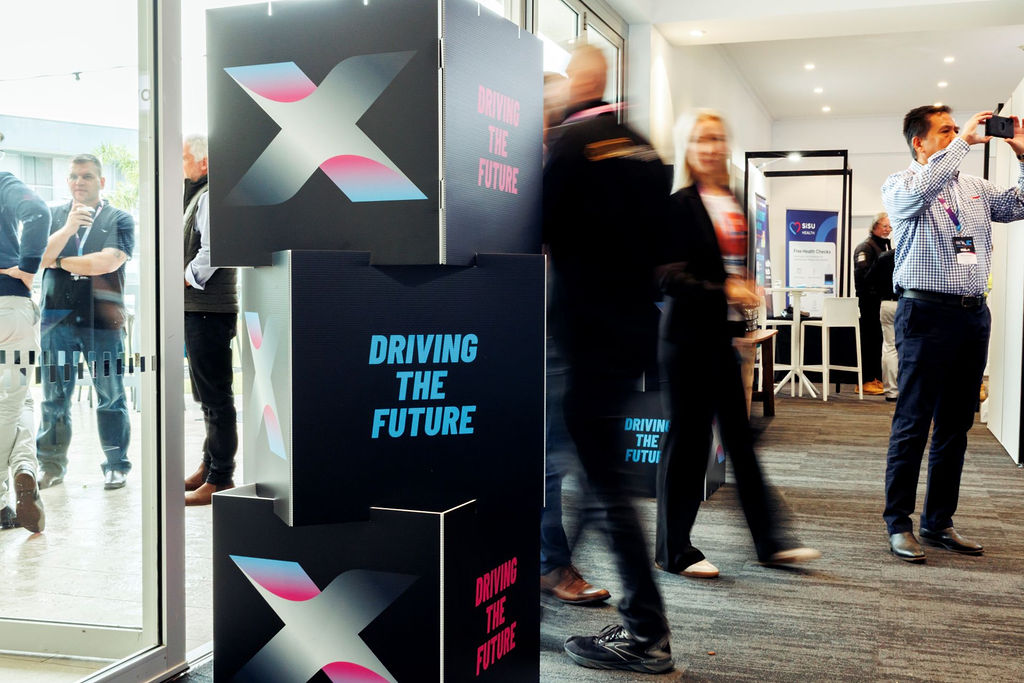 Event lobby with stacked display blocks featuring an X logo and 'DRIVING THE FUTURE' text as guests mingle in the background.