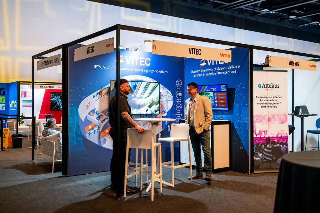 Two men talking at a VITEC trade show booth with a blue graphic backdrop and a large display screen nearby.