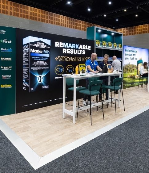 Trade show booth promoting vitamin B products with a bold 'Remarkable Results' display and staff at a white high counter with green stools.