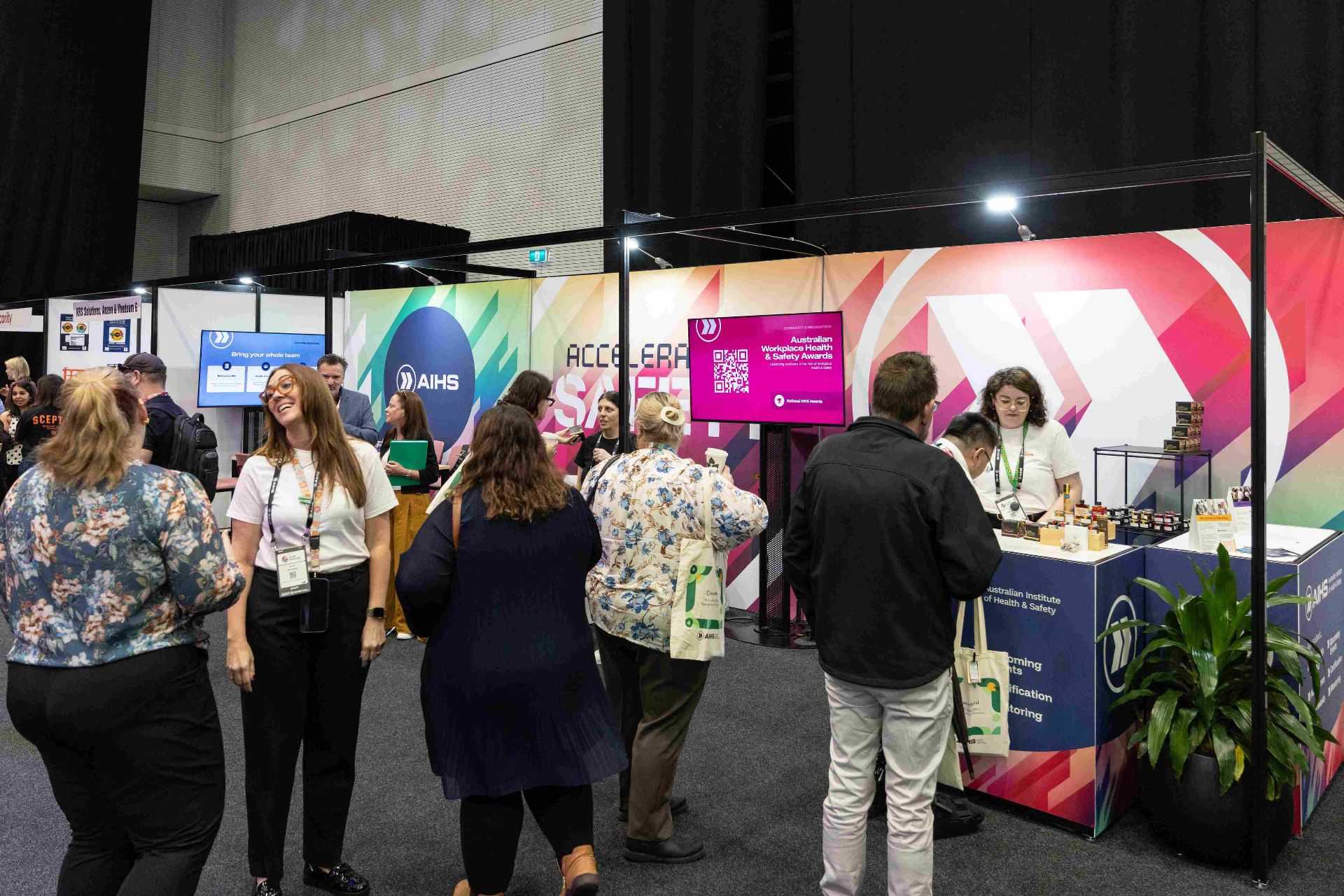 Busy exhibition hall with colorful booth walls, screens, and attendees talking at a trade show stand.