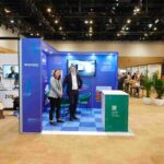 Two professionals stand inside a blue exhibition booth at a trade show, smiling toward the camera.