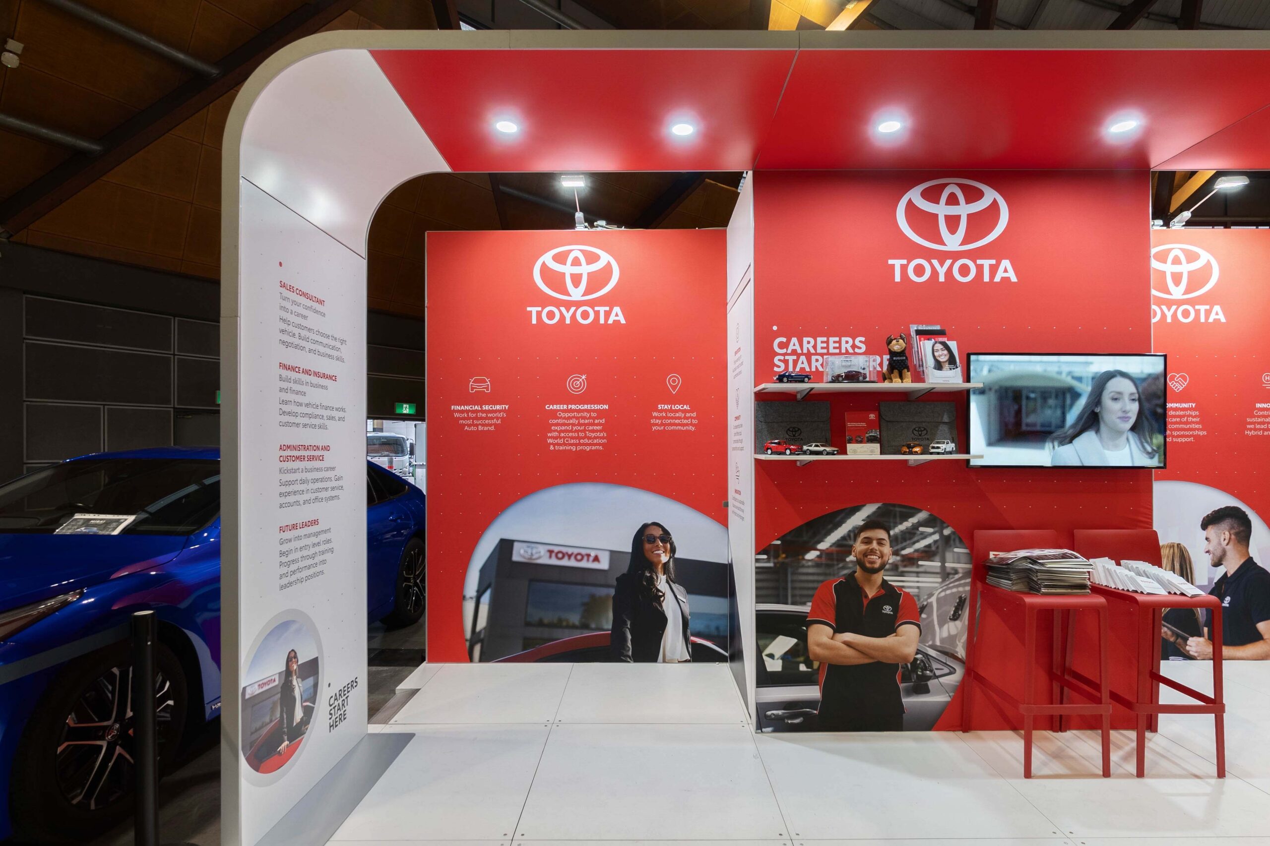 Toyota exhibition booth with red panels, large Toyota logos, and people standing by cars and displays in a showroom setting.