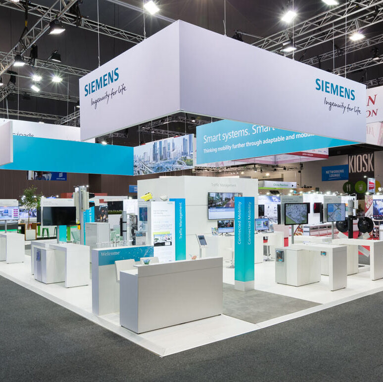 Siemens trade-show booth with white and teal displays, large Siemens banners overhead in a convention hall.