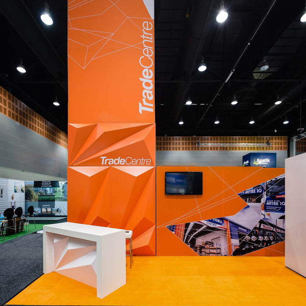 TradeCentre exhibition booth with tall orange geometric backdrop, white counter, and a mounted TV screen in an orange graphic wall scene.