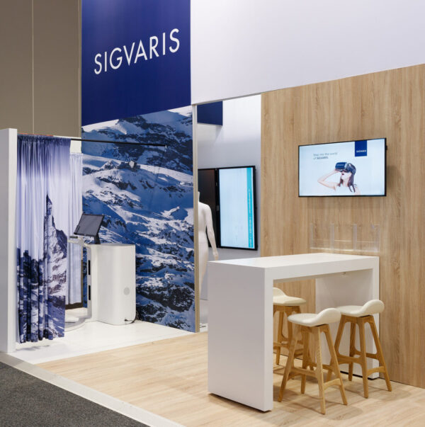 SIGVARIS trade show booth with a blue mountain backdrop, white furnishings, and a touchscreen kiosk in front of a wooden wall.