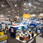 Large indoor trade show hall filled with numerous exhibitor booths, banners, and attendees bustling through aisles. A suspended orange Proxa sign hangs above the crowd.
