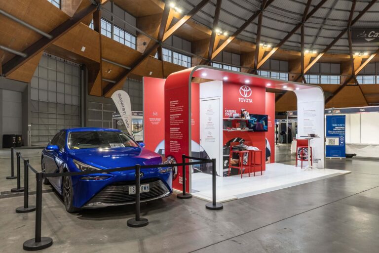 Toyota exhibition booth with red panels and a blue car on display inside a large, well-lit hall.