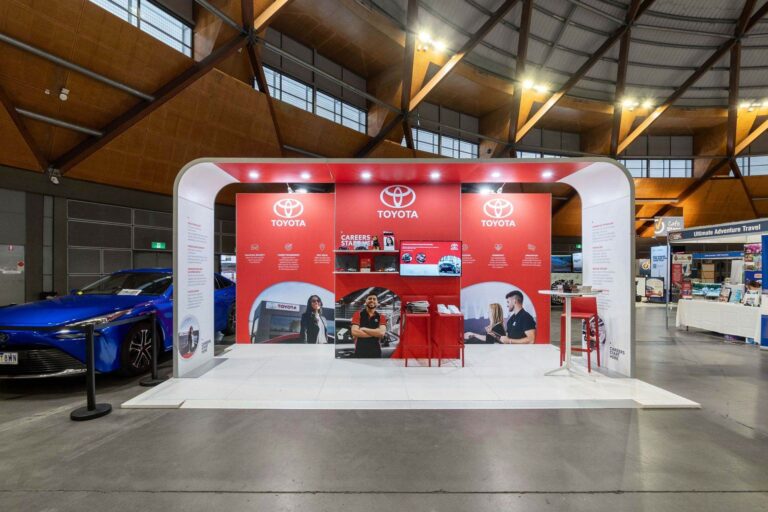 Toyota exhibition booth with red panels and large Toyota logos, a blue car on the left in an indoor convention hall.