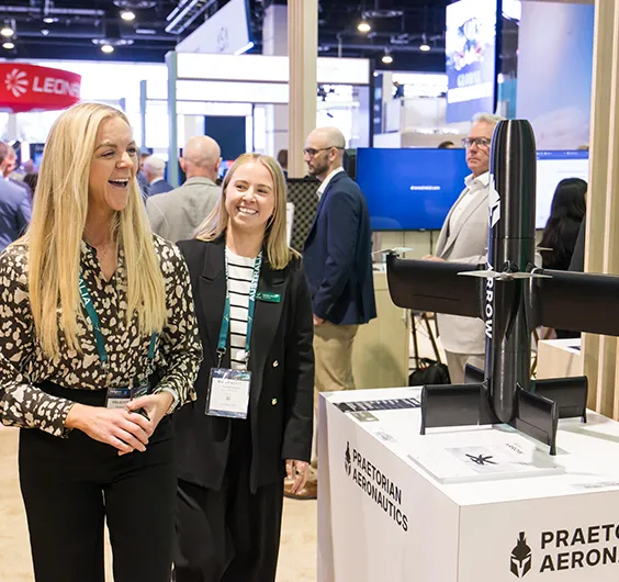 Two smiling women with conference badges at an aviation trade show booth, with a large black propeller model and Praetorian Aeronautics branding nearby.
