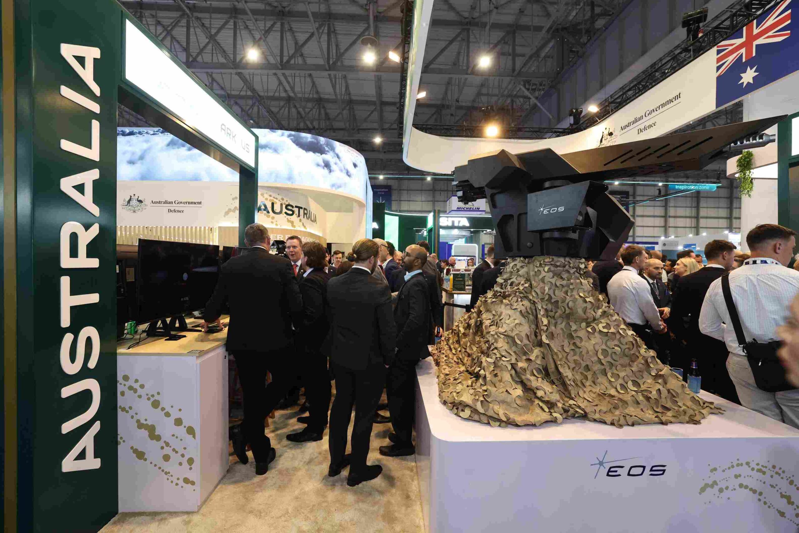 Busy exhibition hall at a defense trade show with Australian Government branding, a tall green pillar reading AUSTRALIA, and a large camouflaged sculpture surrounded by suited attendees.