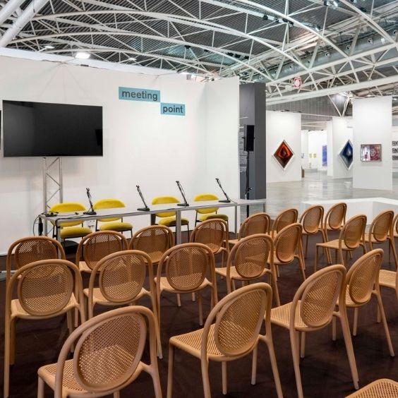 Exhibition hall with a meeting area: rows of wooden chairs facing a panel table with microphones and a large screen.